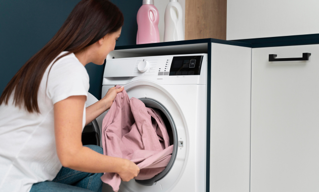 a woman wearing white t-shirt and blue pant is putting clothes in to the washing machine