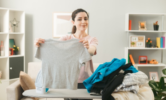 A girl is standing in her room in front of a ironing stand and she is holding agrey t shirt