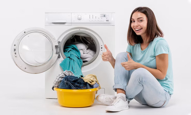 a happy girl sitting on the floor with a bucket full of clothes and a washing machine