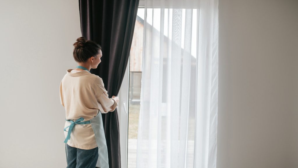 young woman standing near the window and fixing the curtains